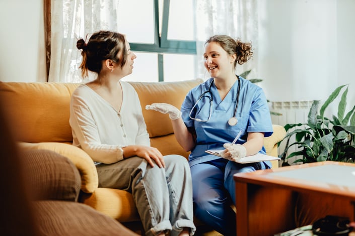 A nurse in scrubs listens and takes notes while talking with a resident on a couch in a home setting, illustrating the relational capabilities—composure, emotional intelligence, attention to detail—that a job title can't capture