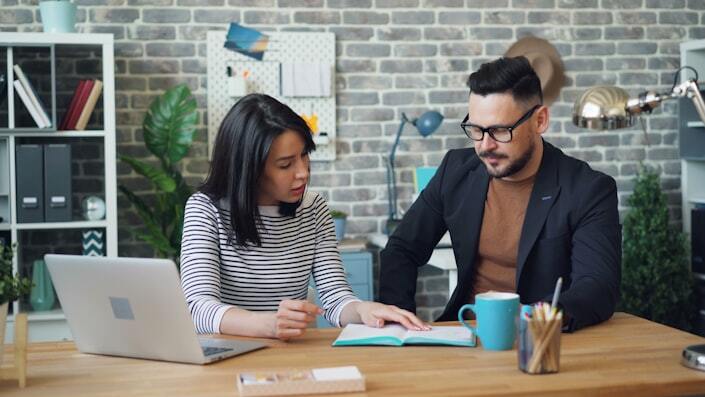 Two coworkers talk through work together at an open-plan office desk