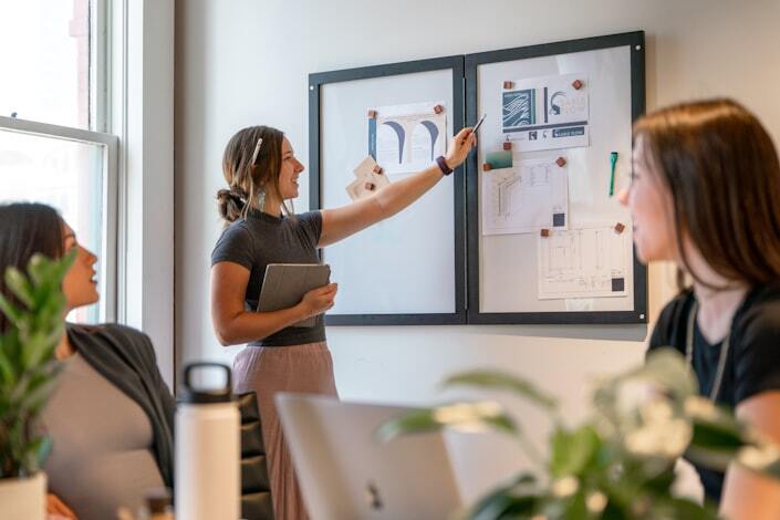 Two women shaking hands across a desk in a modern office setting. The woman facing the camera wears a white blazer, glasses, and a warm smile, with a clipboard in front of her. The other woman, seen from behind, wears a dark blazer. The scene suggests a job interview.