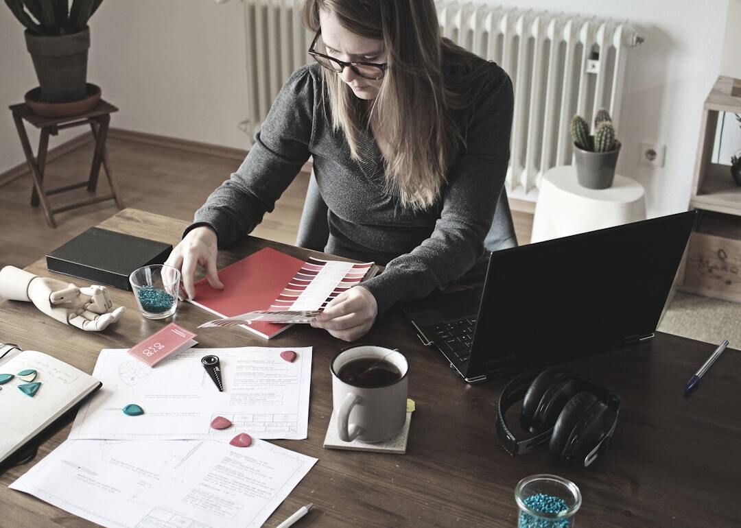 A woman in a dark gray sweater and glasses sits at a wooden desk in a home office, examining color swatches in shades of red and pink. The desk is covered with design materials.  A laptop, a mug of coffee, and black over-ear headphones sit to her right. A radiator, potted cacti, and a houseplant are visible in the background. The scene suggests a creative professional working from home.